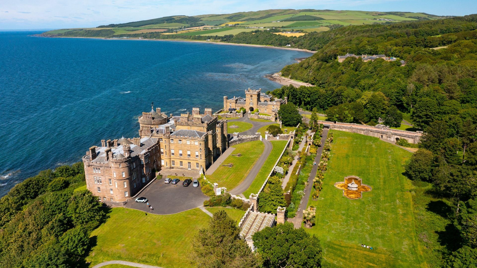 An aerial view of a large stone castle on a lush green cliffside overlooking the ocean.