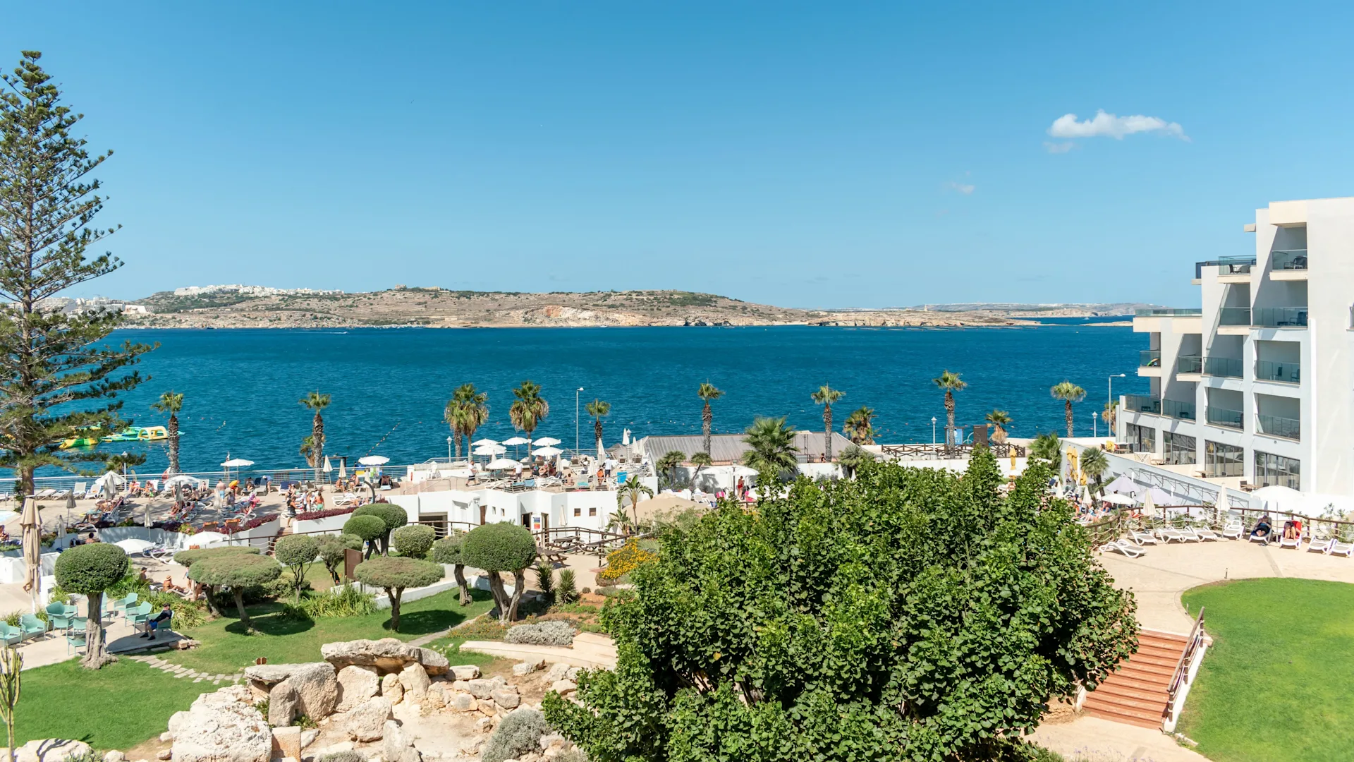 A sunny view of a coastal resort with white buildings and lush greenery overlooking the sea.