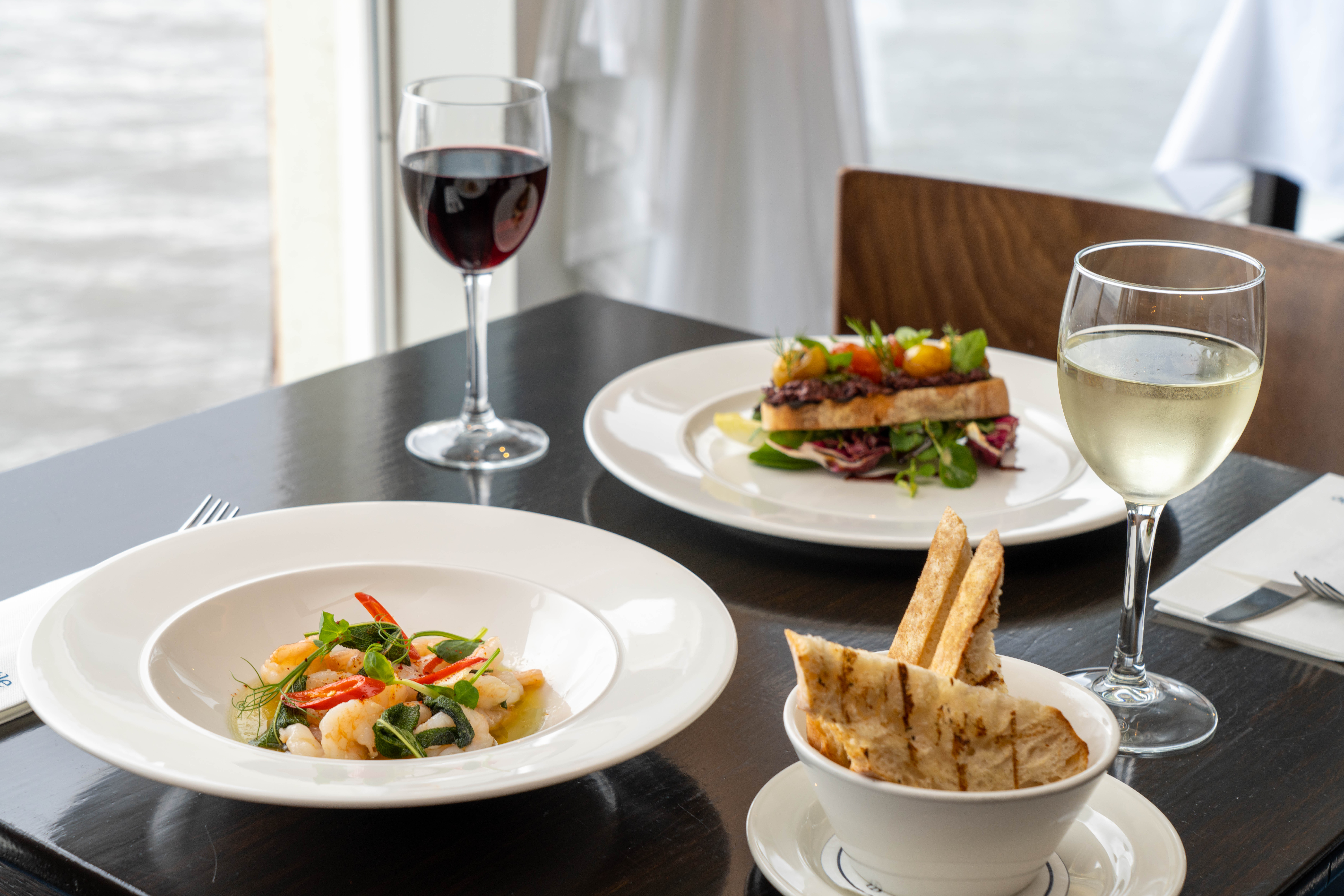Two plated meals with red and white wine on a dark wooden table by a window.