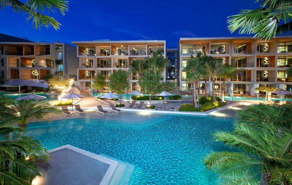 A modern resort hotel with a large, illuminated swimming pool and palm trees at night.