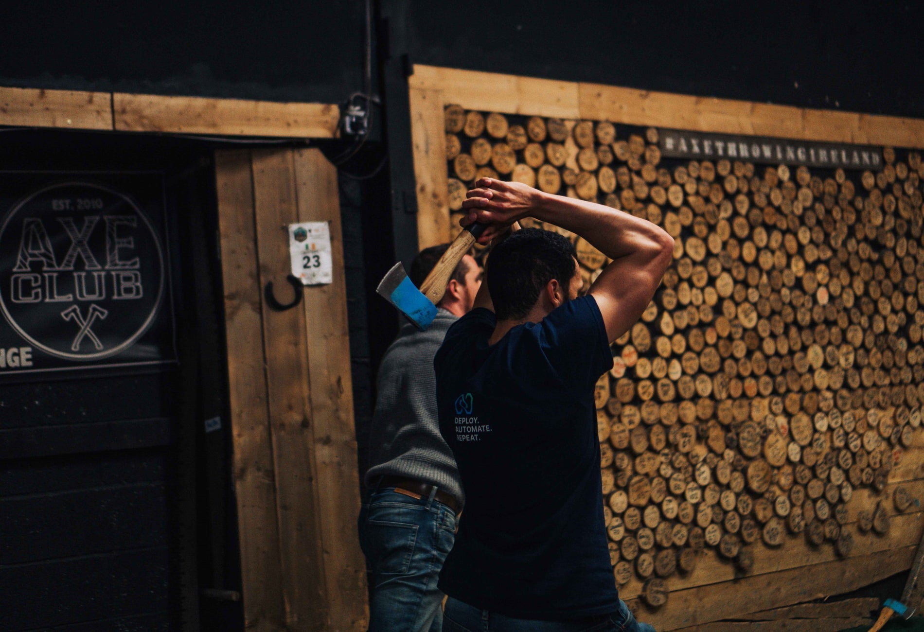 A man seen from behind holds an axe over his head, about to throw it at an axe club.