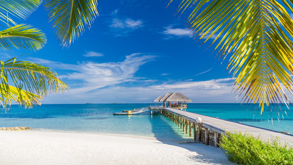 A wooden pier with a thatched-roof hut extends over turquoise water from a white sand beach.