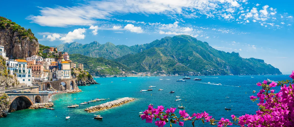 A colorful town built into a seaside cliff, overlooking a turquoise bay with boats and mountains.