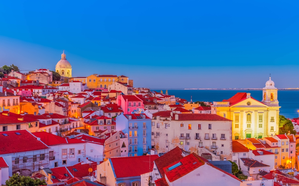 A vibrant cityscape with colorful, red-roofed buildings overlooking the water at twilight, with a large dome visible.