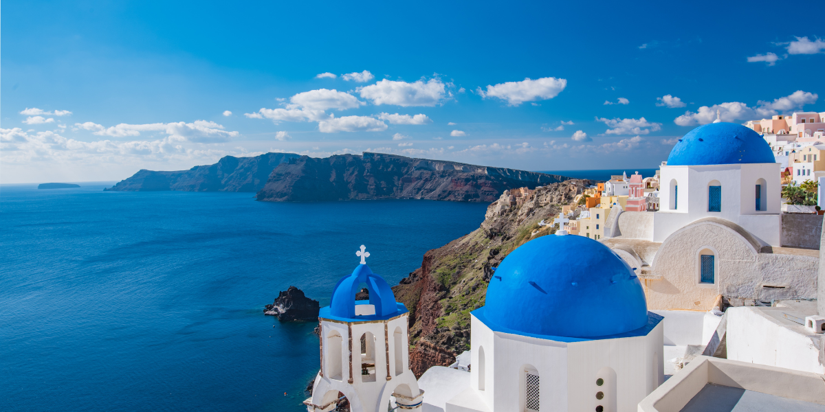 White buildings with blue domes overlooking a sea and distant cliffs.