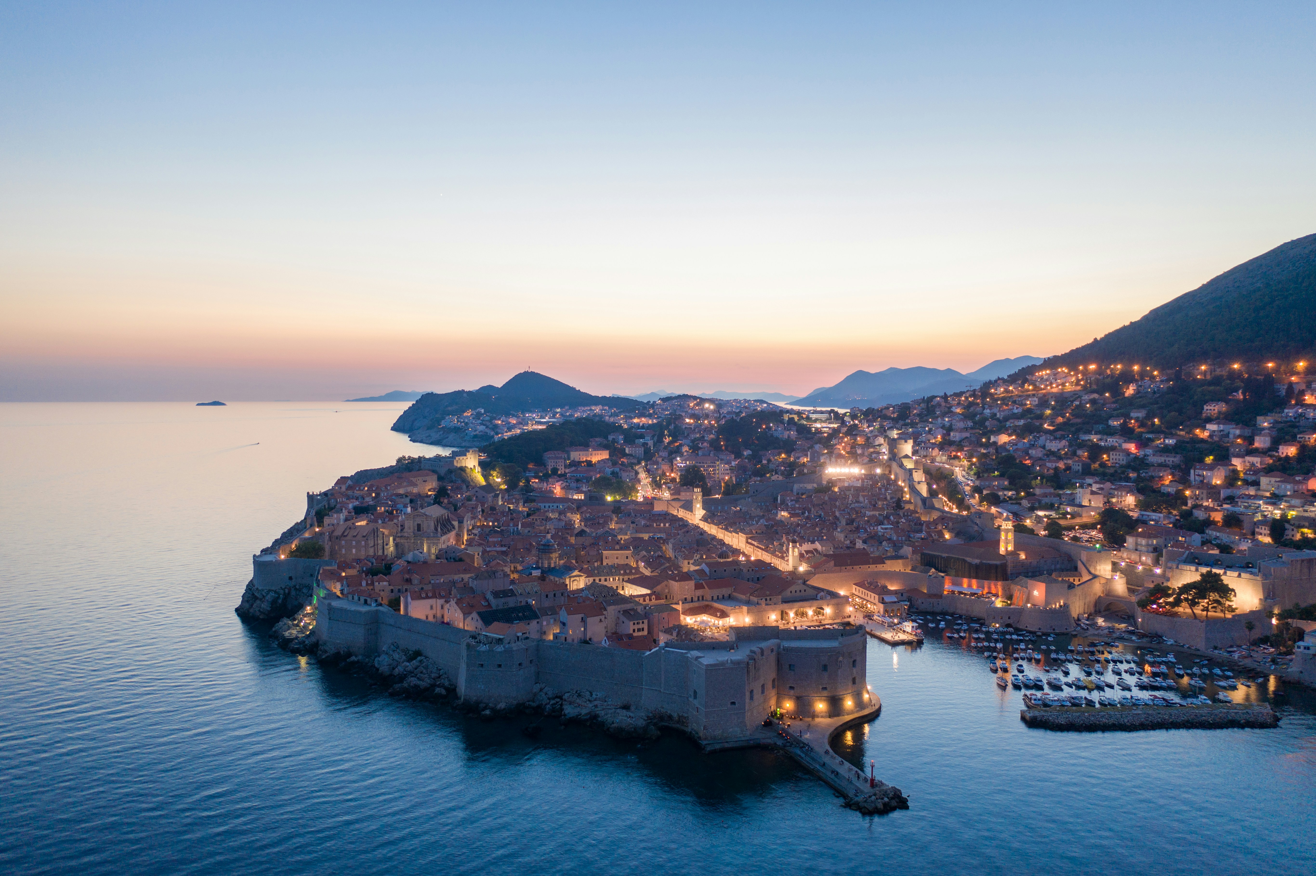 Aerial view of a coastal city at sunset with illuminated buildings and surrounding hills.