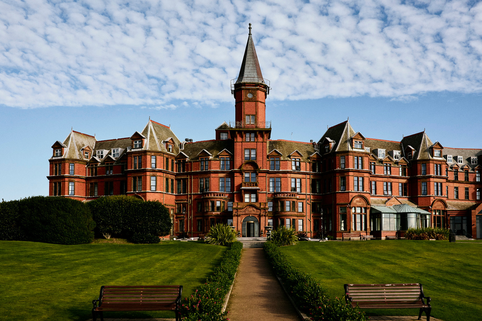 A large, red-brick building with a central tower, viewed from a path across a manicured green lawn.
