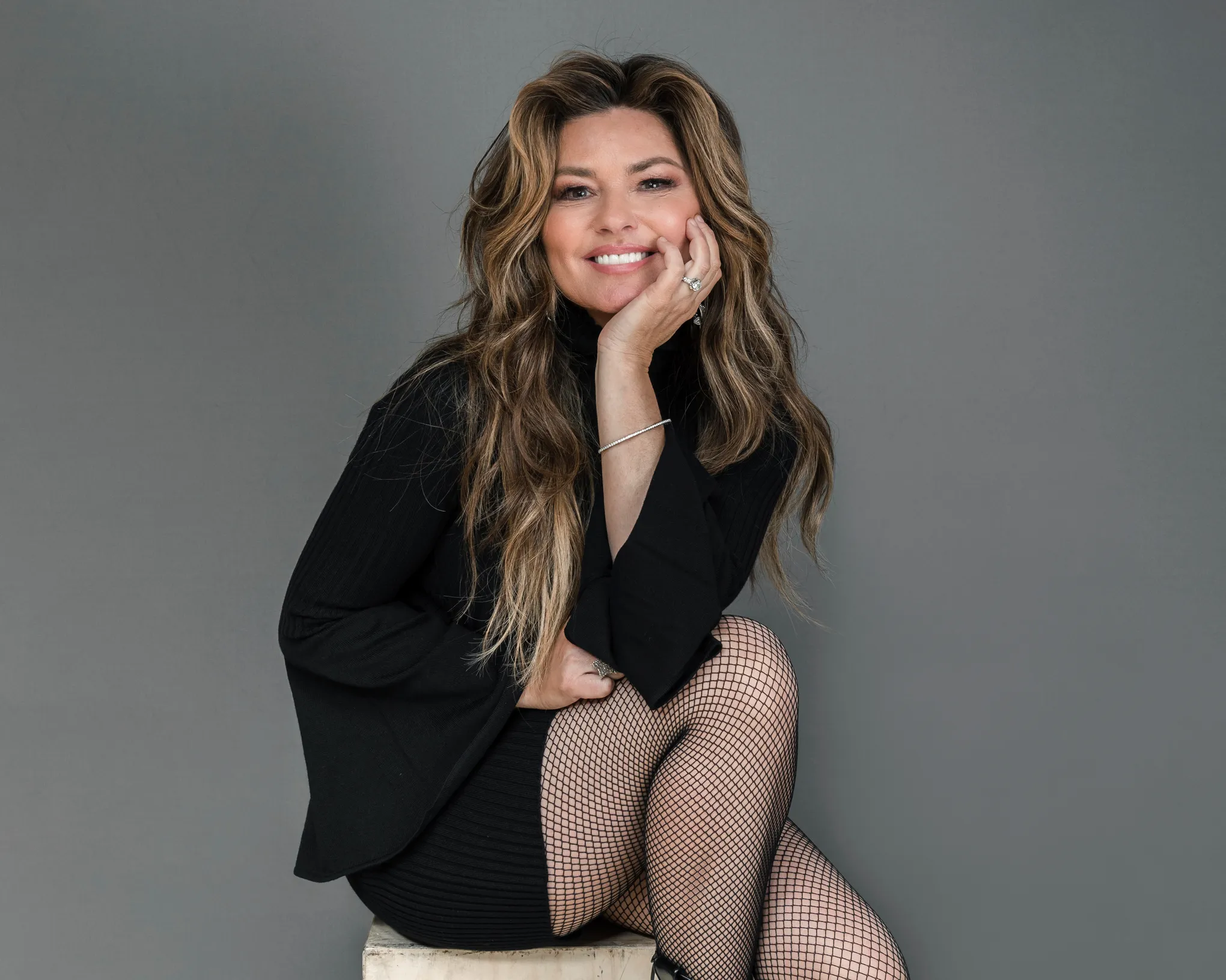 Shania Twain poses in a black turtleneck and fishnet stockings against a gray studio backdrop.