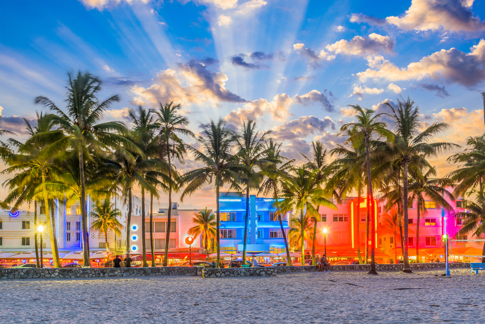 Palm trees on a beach in front of colorful, neon-lit buildings under a dramatic, cloudy sky at dusk.