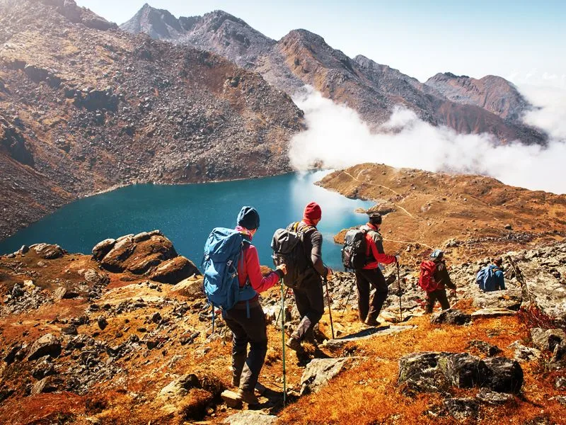 Hikers with backpacks walking down a rocky mountain path near a blue lake.