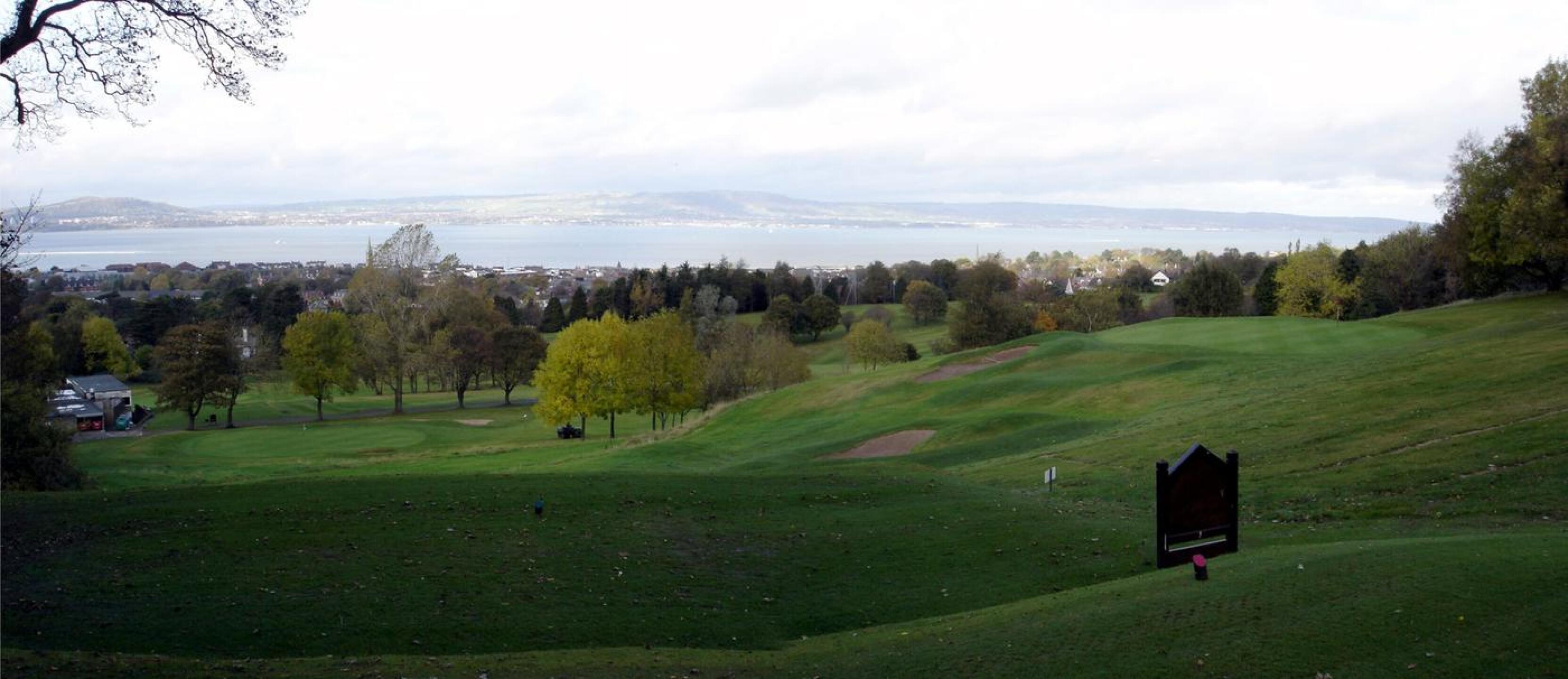 Golf course with trees, overlooking a body of water and distant hills.