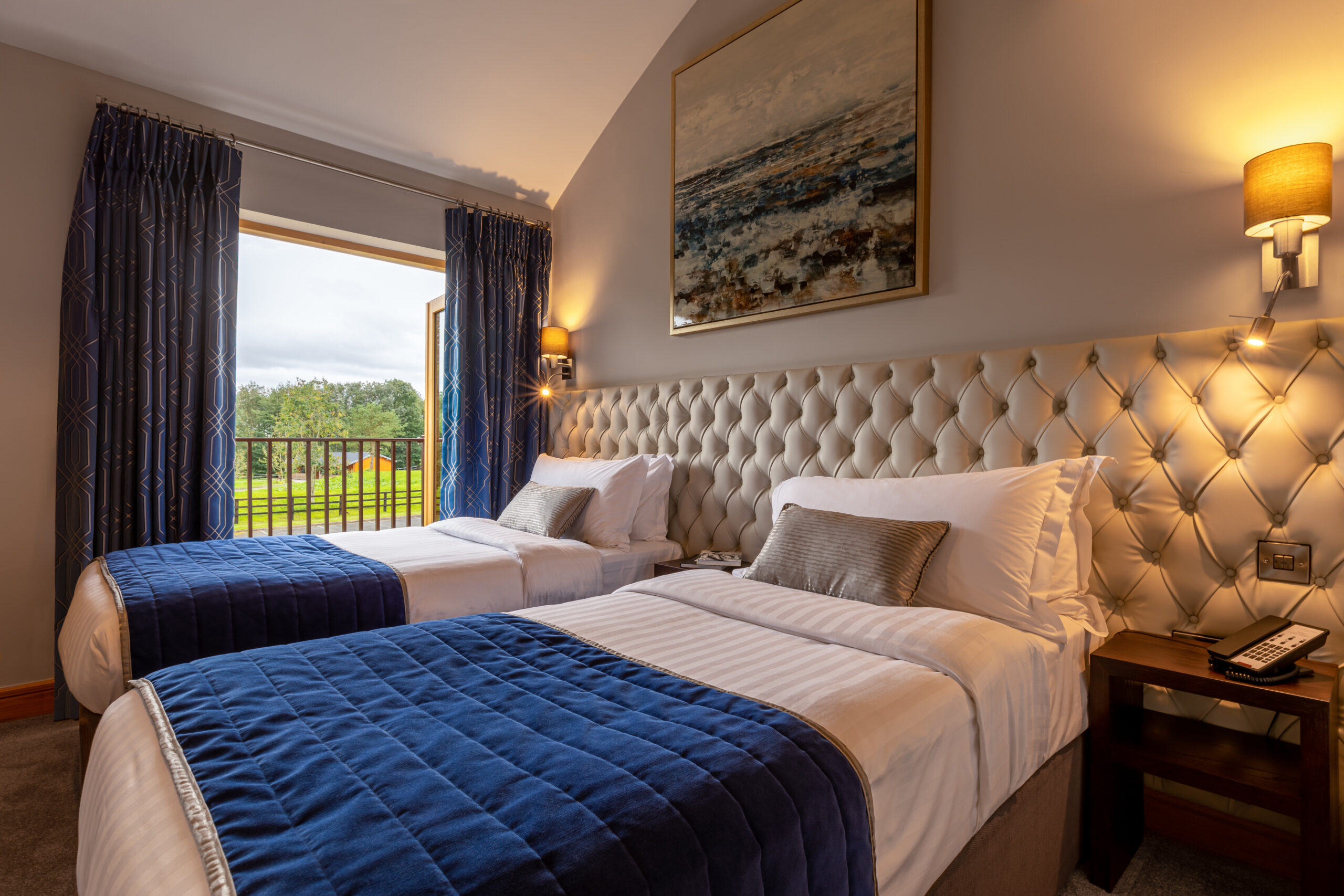 A hotel room with two twin beds with blue throws and an open door leading to a balcony.