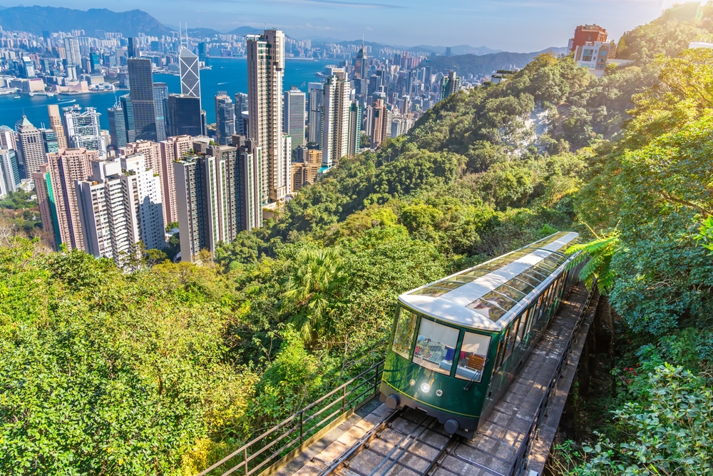 A green funicular ascends a lush hillside overlooking a modern city skyline and harbor.