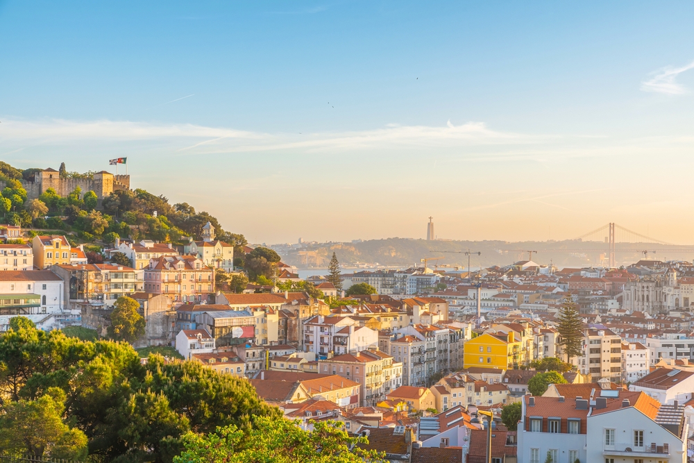 Cityscape with colorful buildings, a castle, and a suspension bridge at sunset.