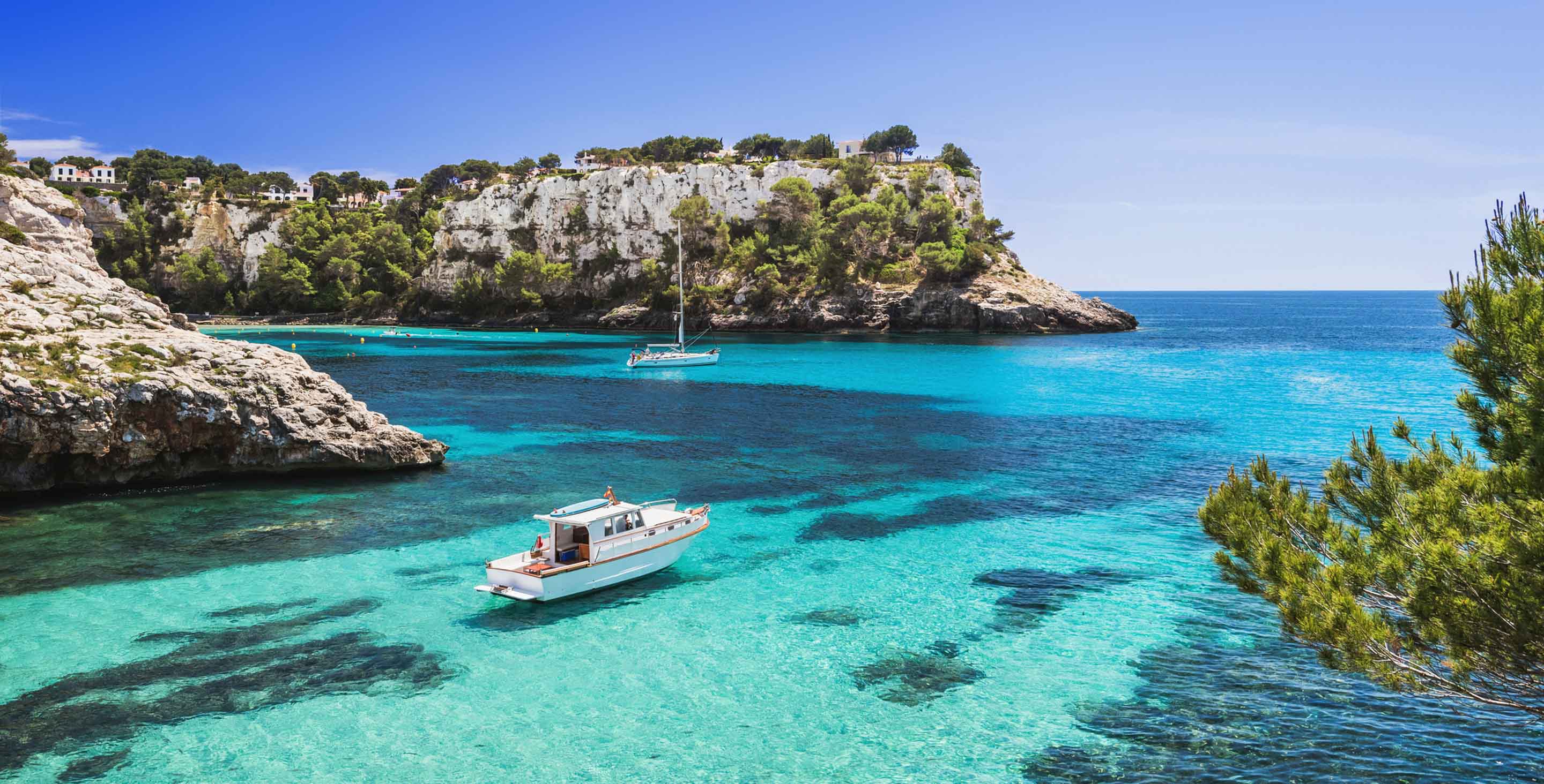 Clear turquoise sea with boats and rocky cliffs under a blue sky.