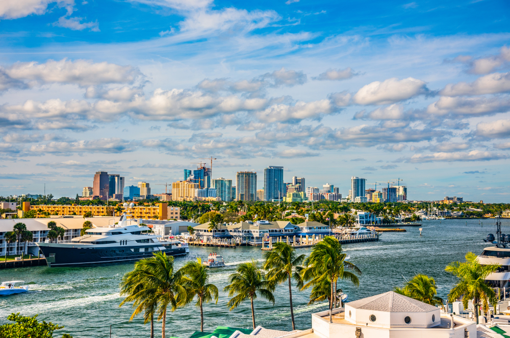 A city skyline with a waterway in the foreground, featuring boats, a large yacht, and palm trees.