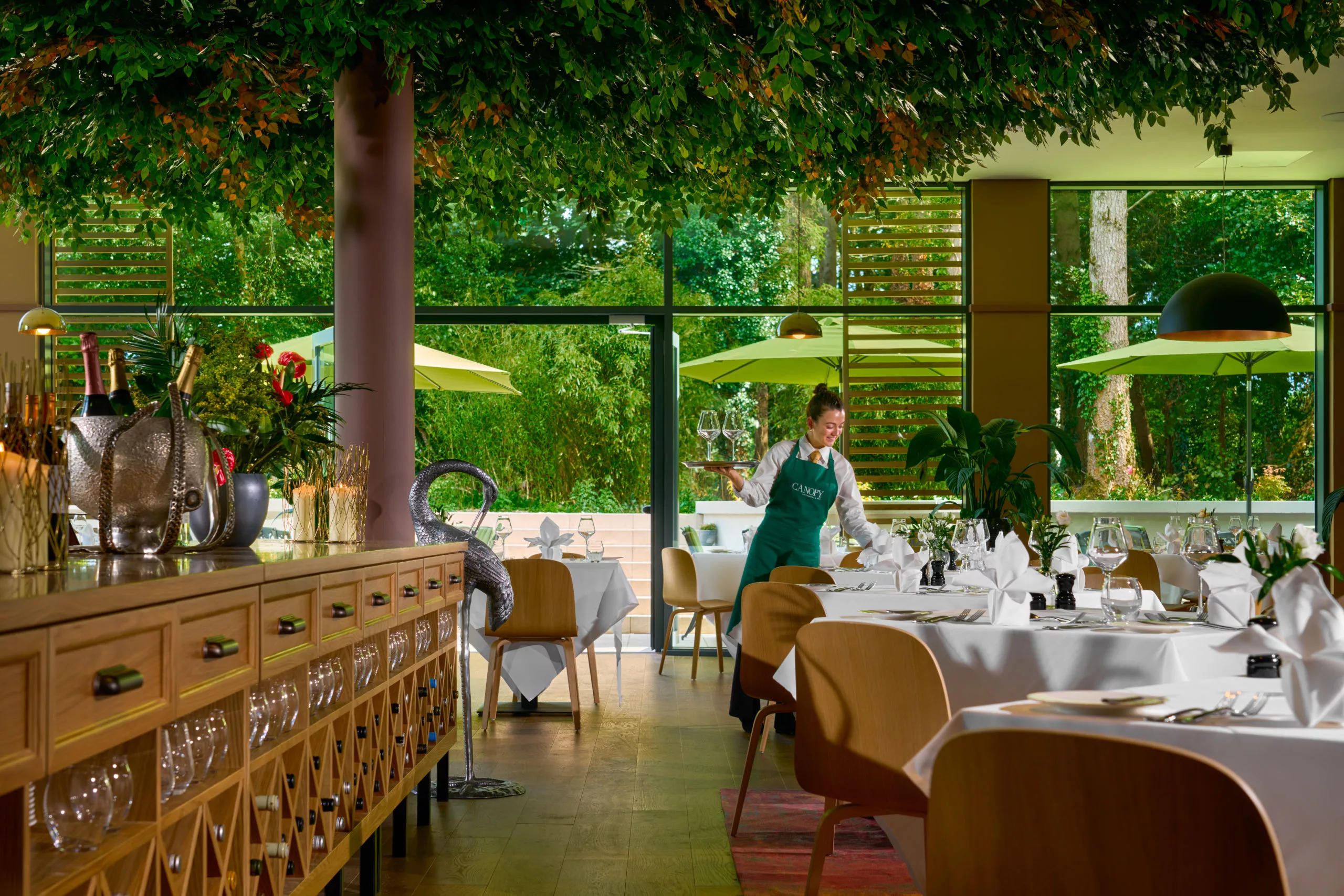 Elegant restaurant with a leafy ceiling, set tables, and a server arranging glasses.