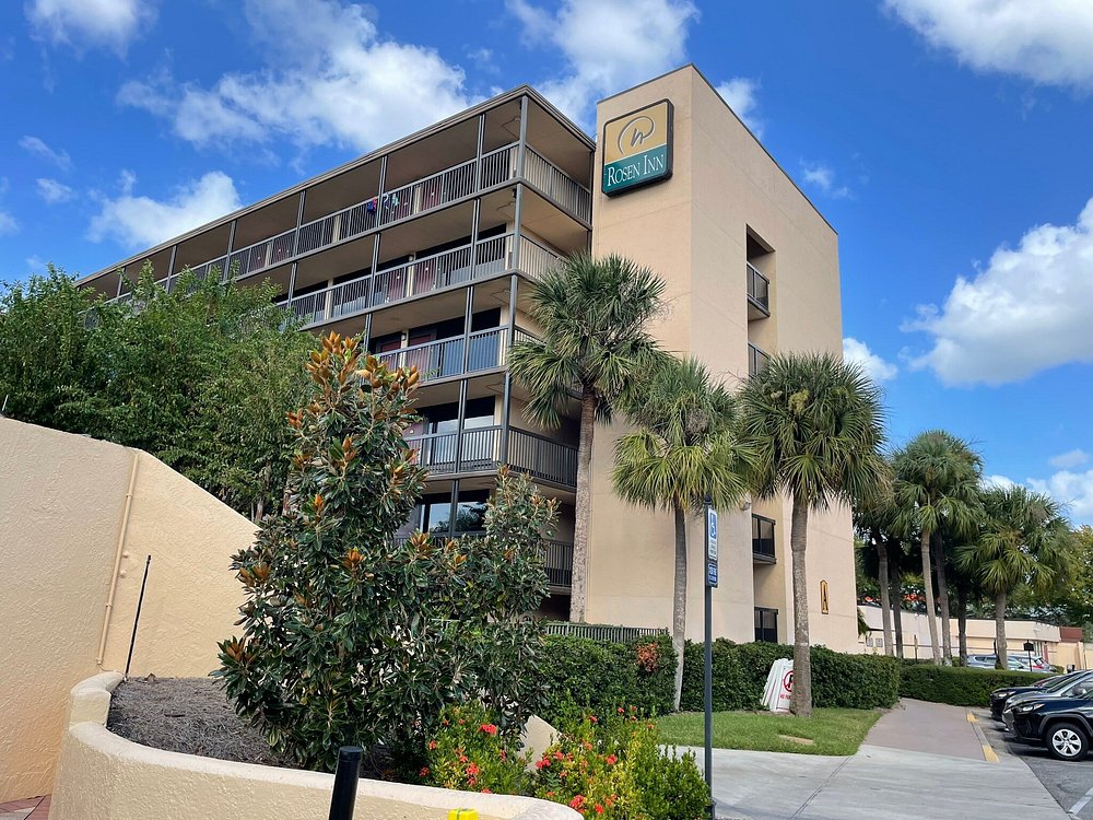 Four-story hotel building with balconies, palm trees, and a sign reading 'Rosen Inn'.