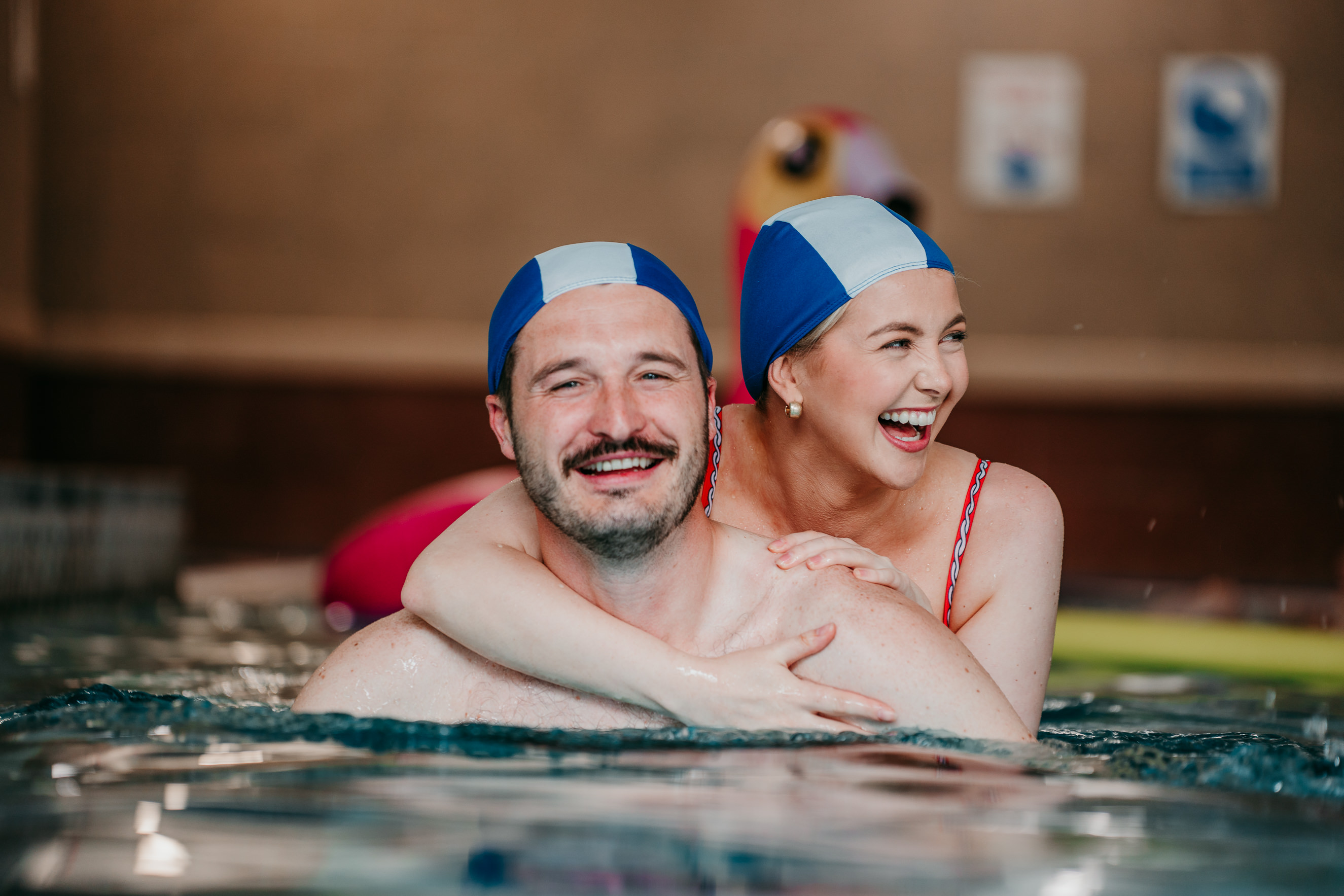 A smiling man and laughing woman wearing matching swimming caps embrace in a pool.
