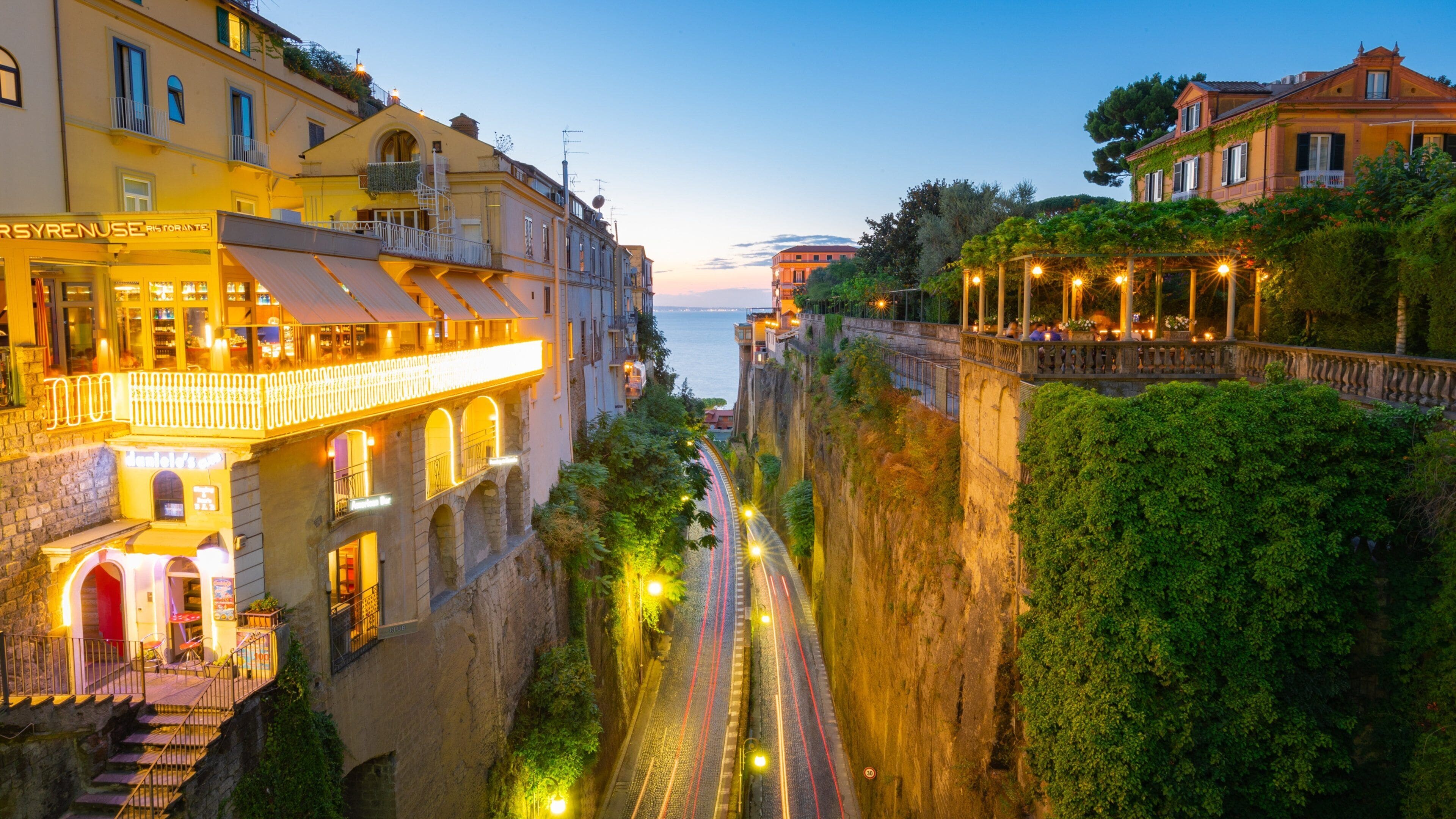 A coastal town's illuminated buildings flank a deep road leading to the sea at dusk.
