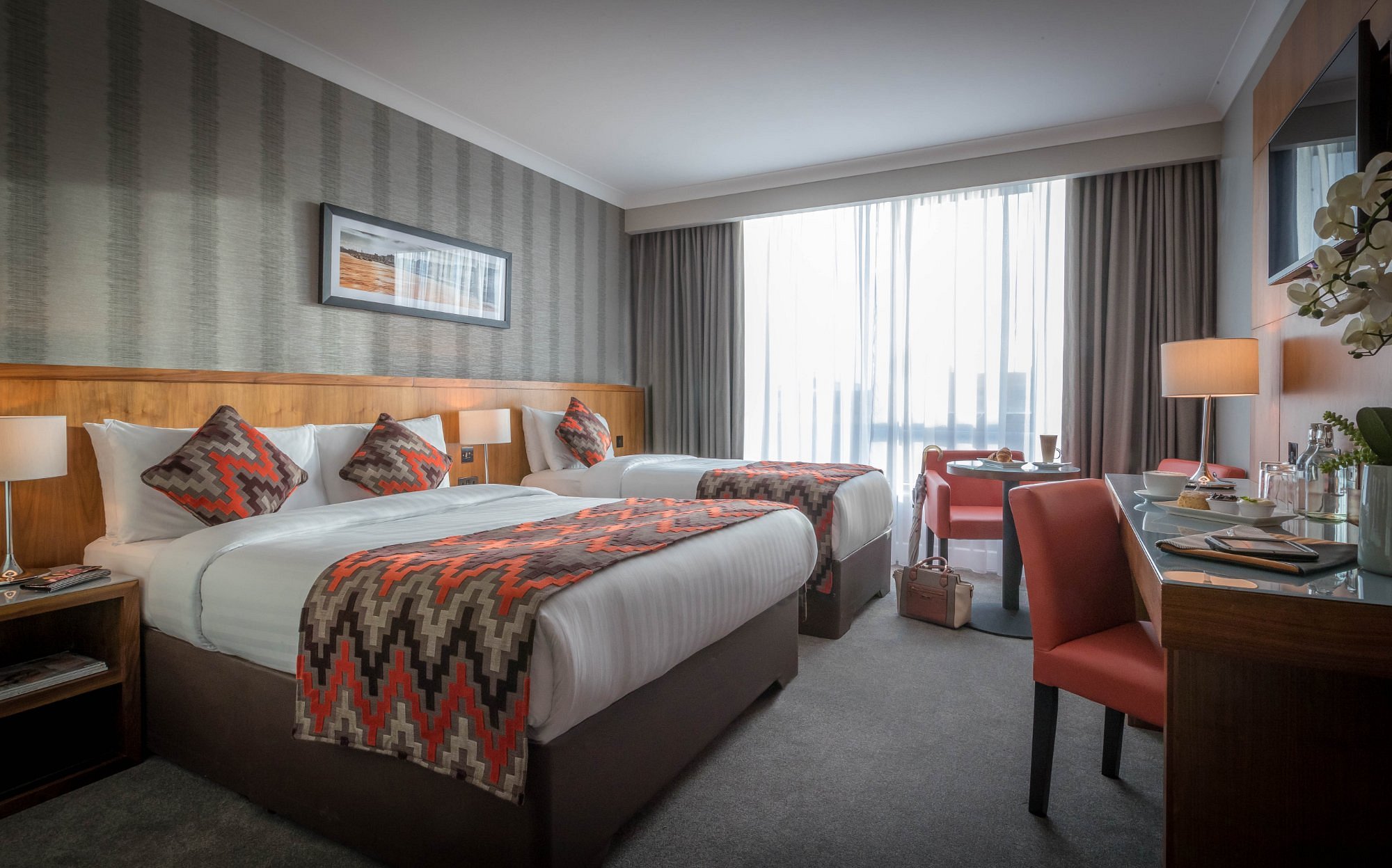 Hotel room with two beds, patterned pillows, desk, chairs, and large window with curtains.
