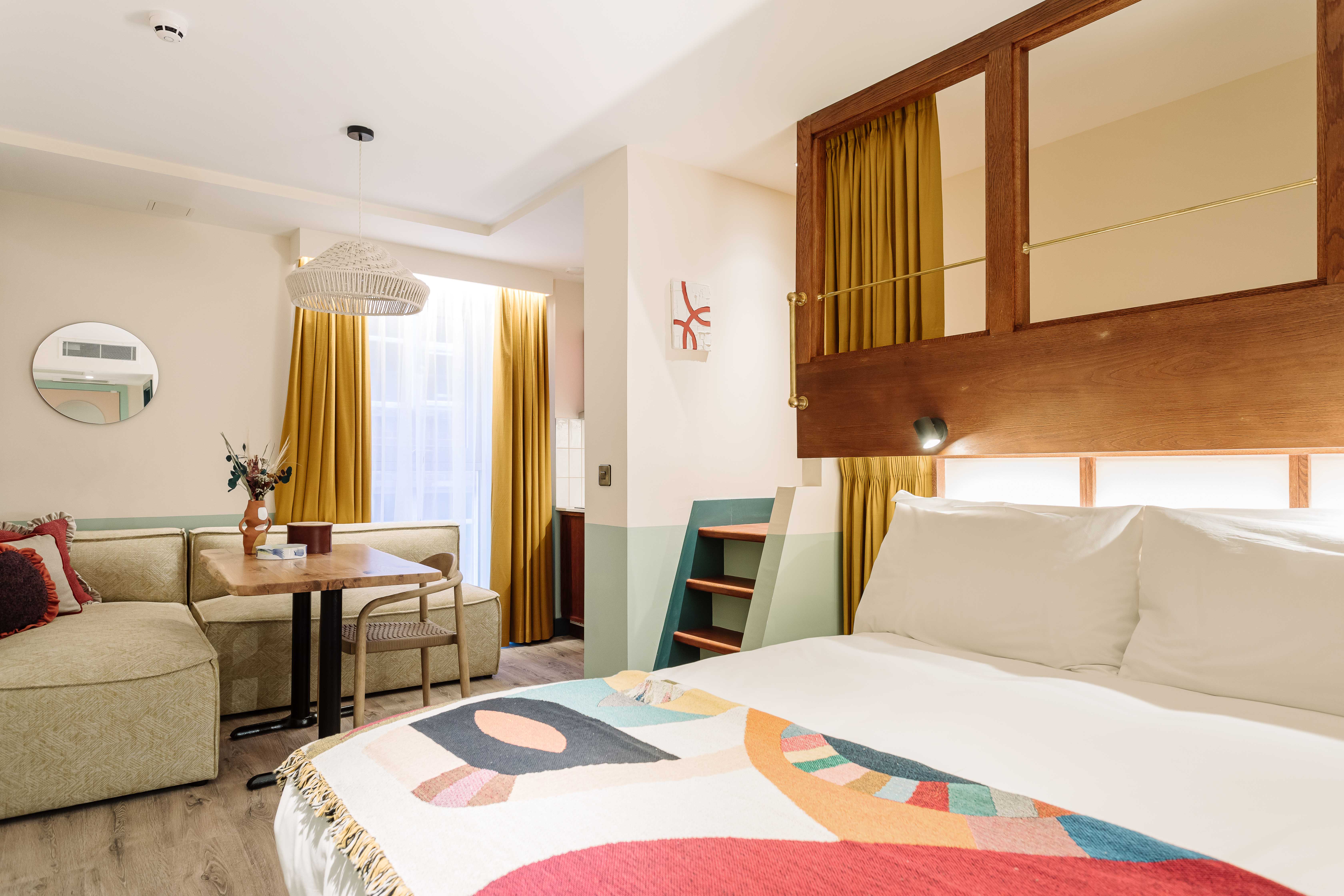 A stylish, well-lit hotel room with a bed, a small seating area, and mustard-colored curtains.