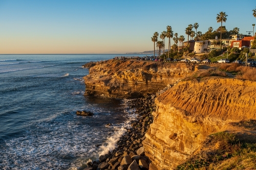 A scenic view of rocky cliffs overlooking the ocean at sunset, with palm trees and buildings.