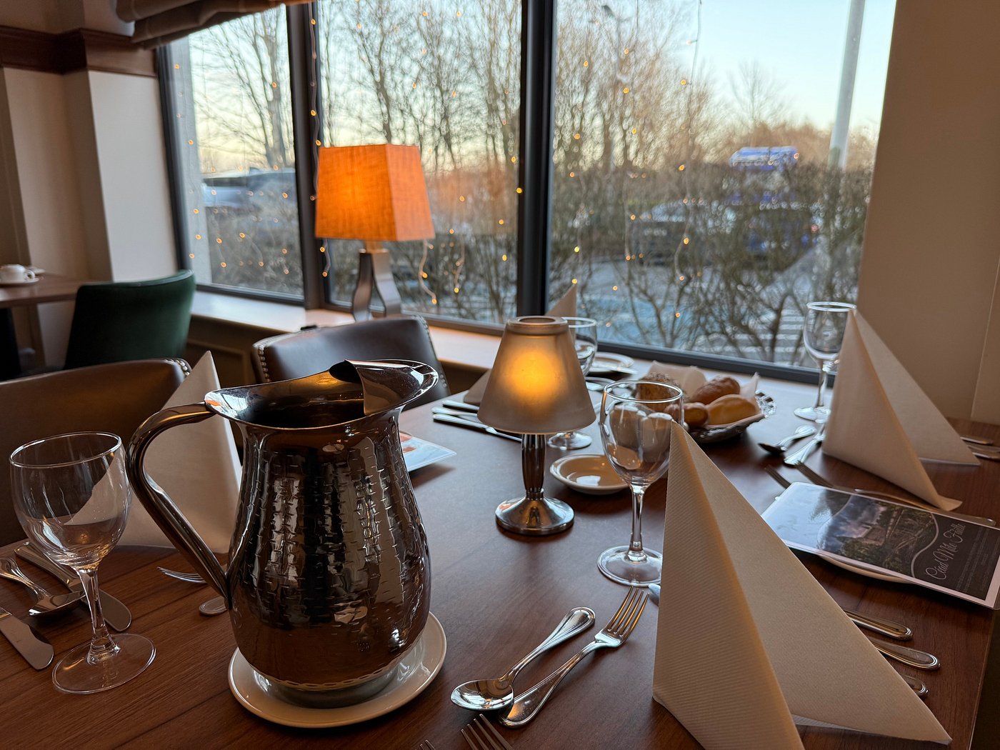 Table set with glasses, napkins, and a metal pitcher by a window with a lamp.