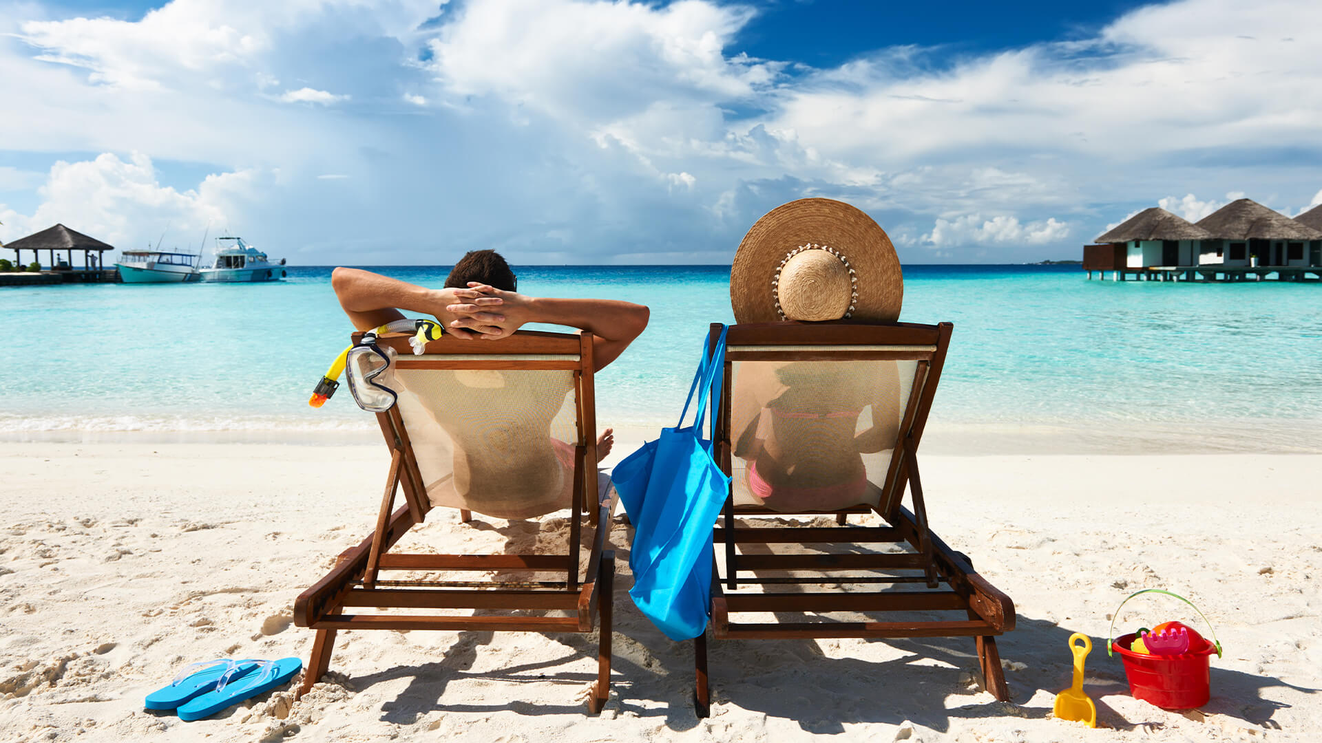 Two people relaxing on beach chairs by the ocean, with boats and huts in the background.