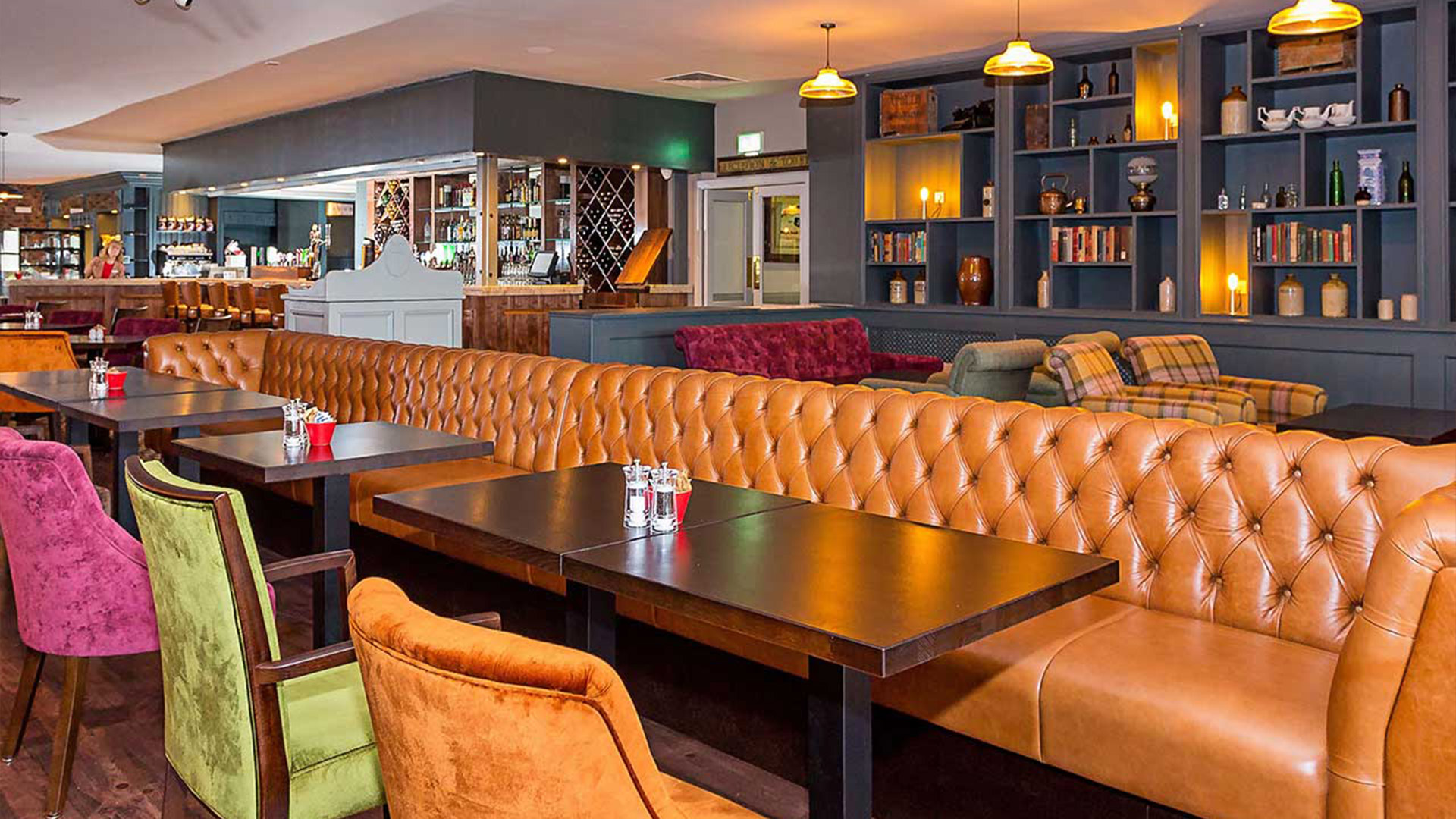 A warmly lit restaurant interior with a long, tufted leather banquette, tables, and colorful chairs.