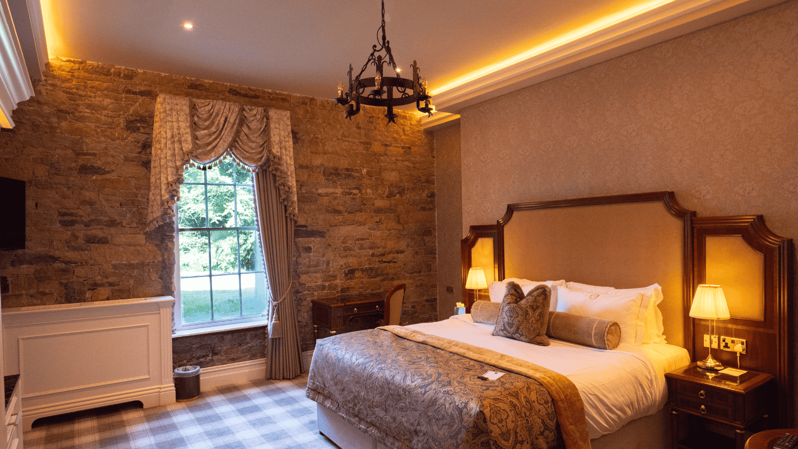 A luxurious hotel room with an exposed stone wall, a large bed with a patterned coverlet, and a chandelier.