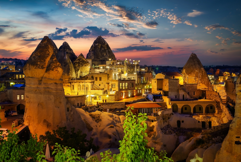 A town built into conical rock formations, illuminated at dusk under a colorful sky.