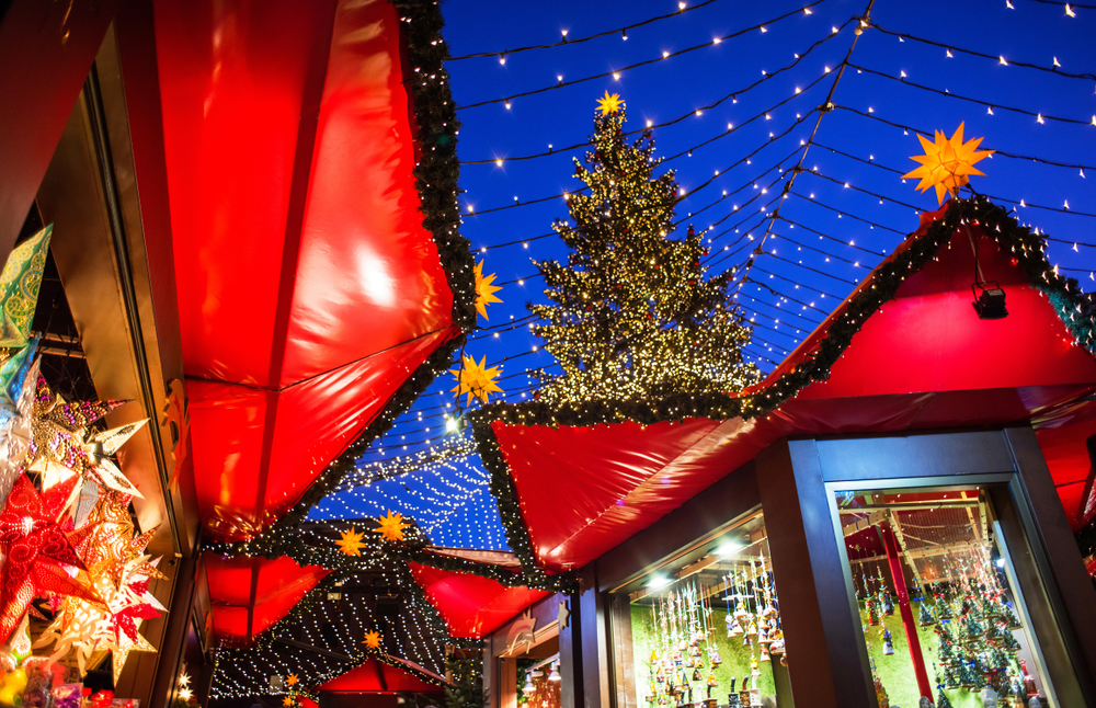 A Christmas market at night, with red-roofed stalls and a large, illuminated Christmas tree under string lights.