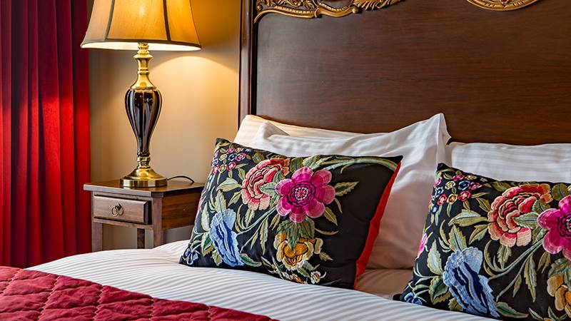 A close-up of a made bed with floral embroidered pillows next to a lit bedside lamp.
