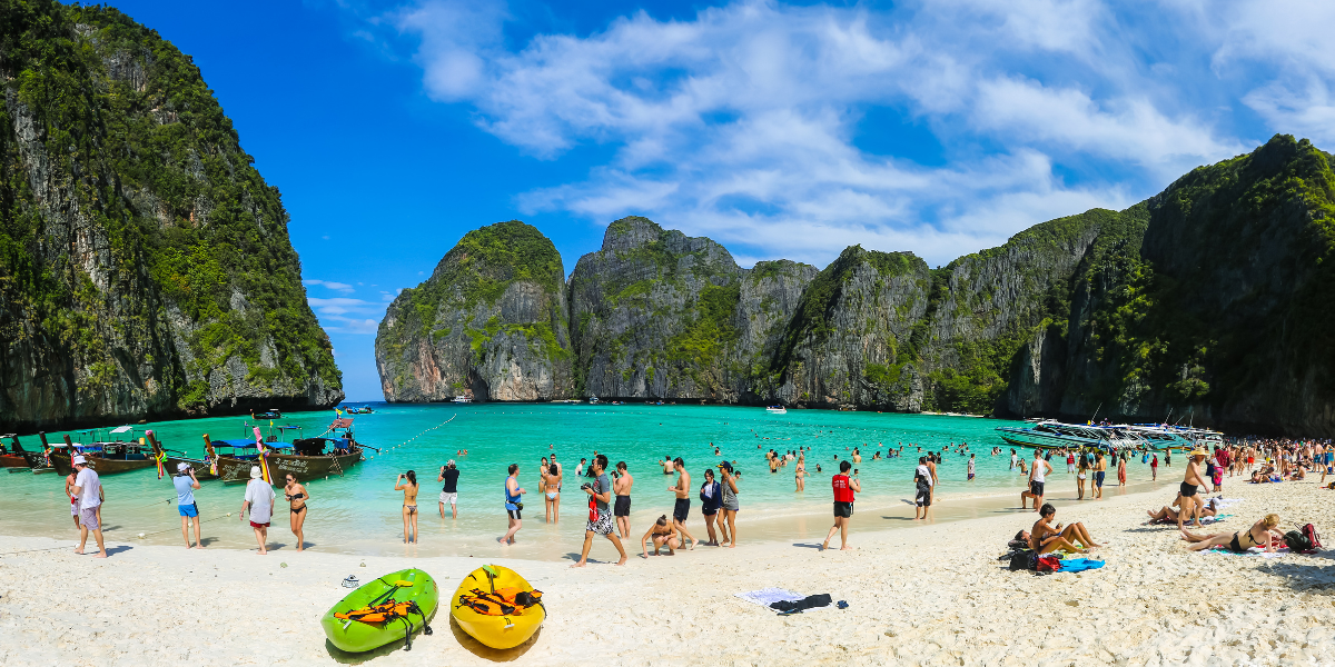 Crowded beach with people swimming, boats, and limestone cliffs in the background.