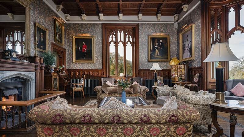 A lavishly decorated, wood-paneled sitting room with a fireplace, patterned sofas, and framed portraits on the walls.