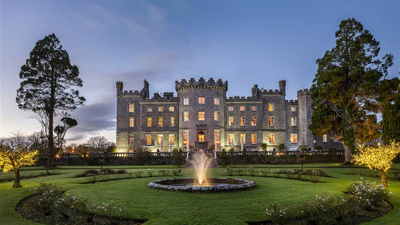 A large stone castle with illuminated windows at dusk, seen across a manicured lawn with a central fountain.