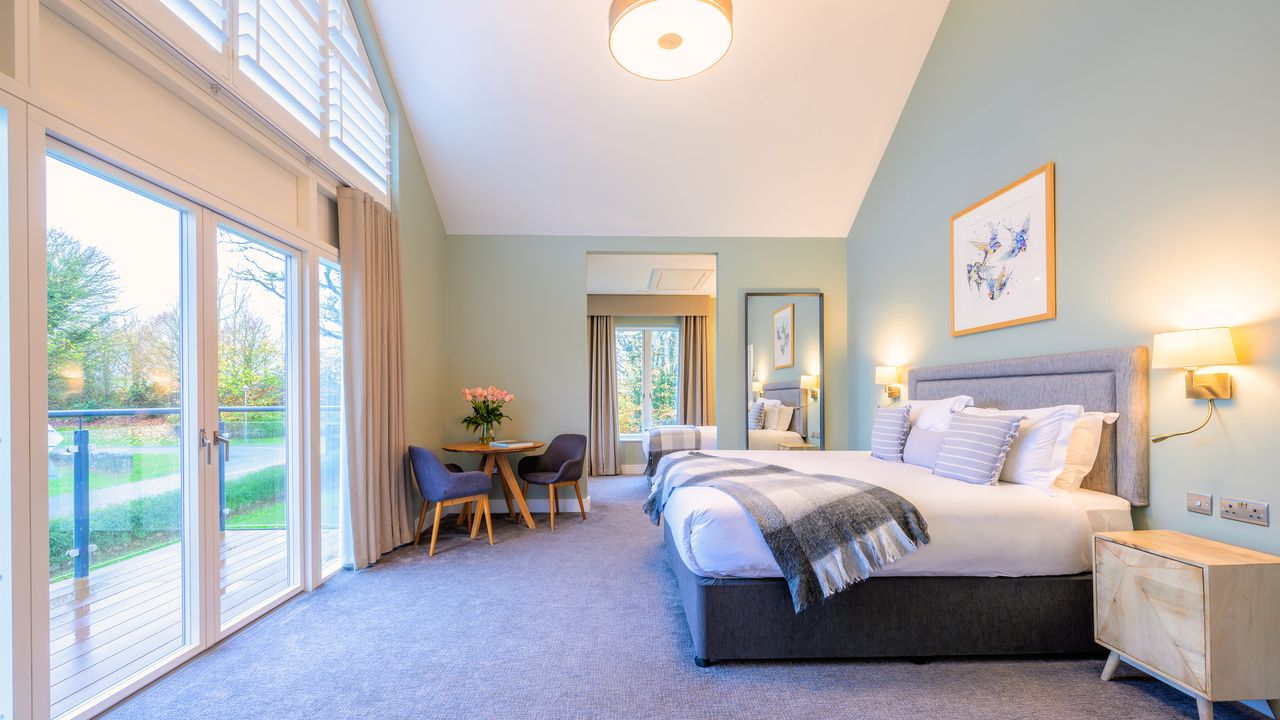 A brightly lit, spacious hotel bedroom with a large bed and glass doors leading out to a balcony.