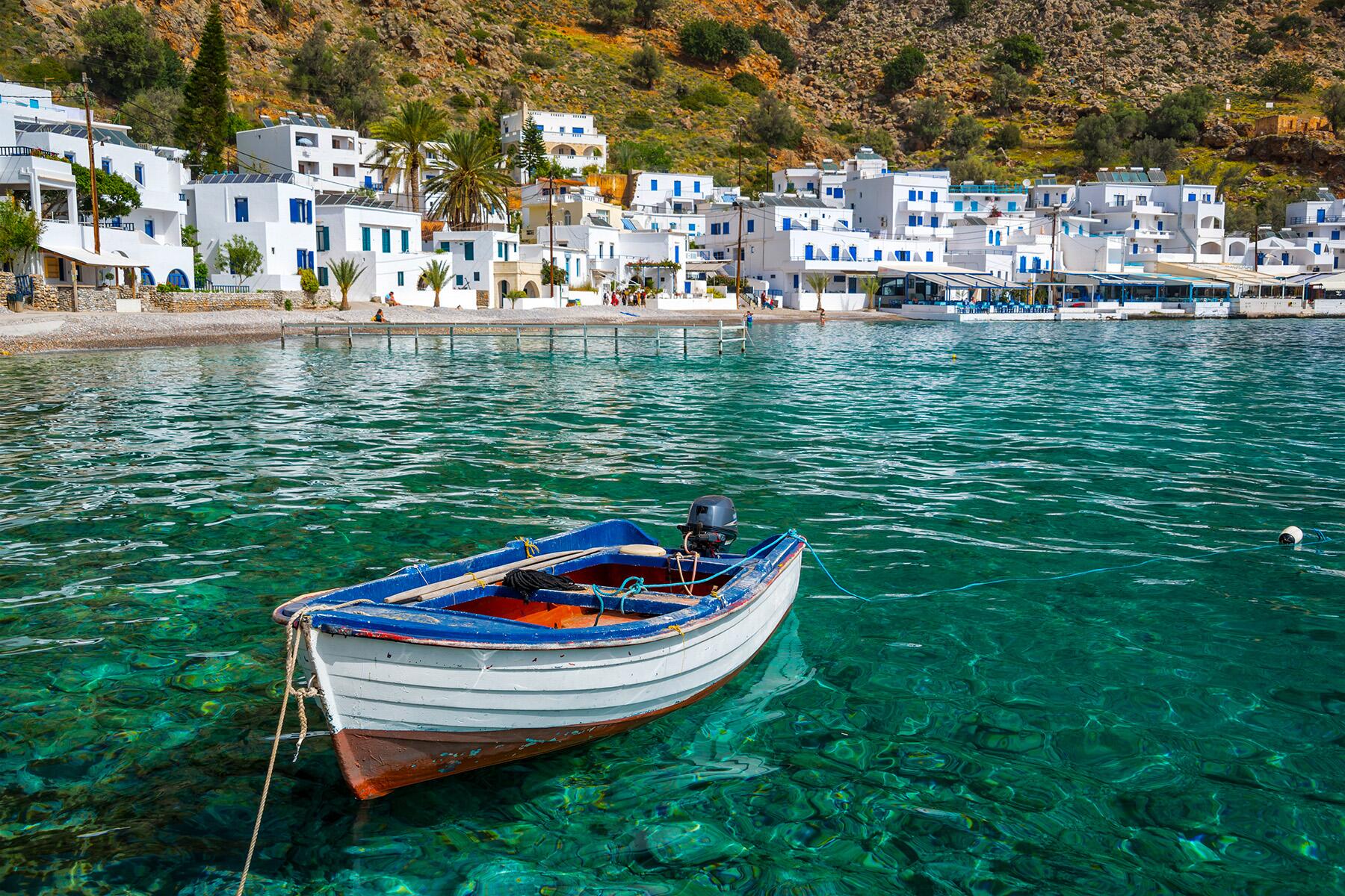 Small boat on clear turquoise water near white buildings on a hillside.