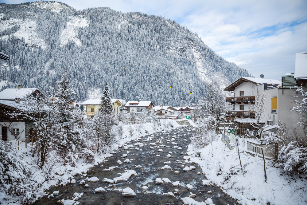 A river flows through a snow-covered village at the base of a large, tree-covered mountain.