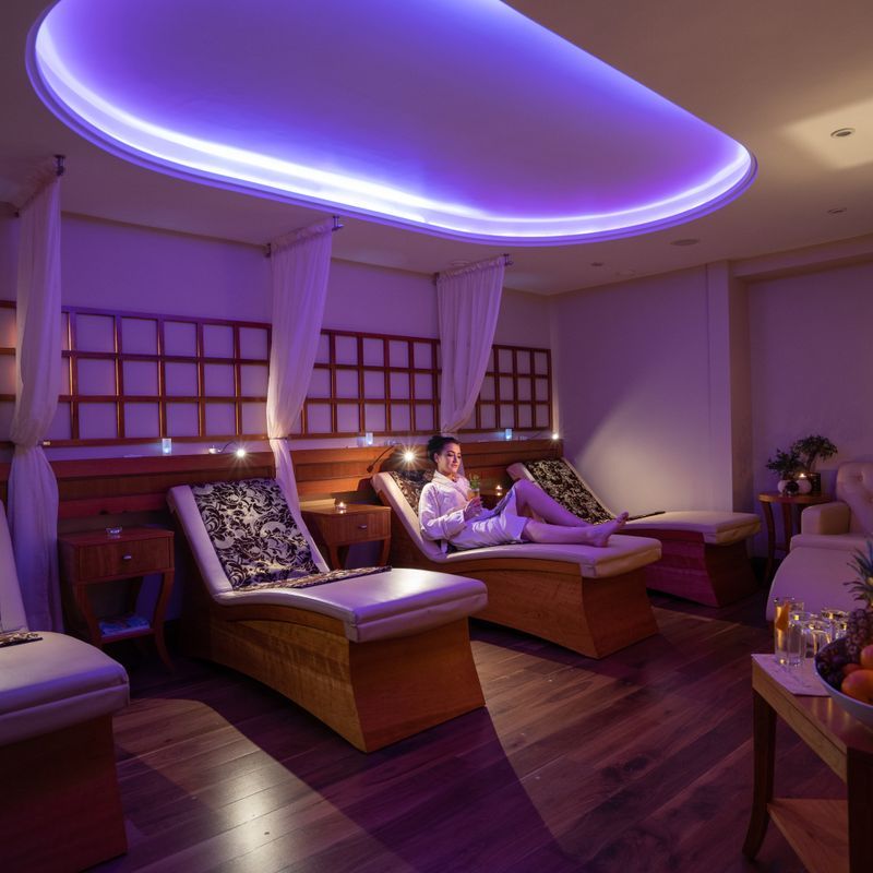 A woman in a white robe relaxes on a lounge chair in a dimly lit spa room.