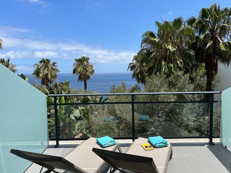 A balcony with two lounge chairs overlooking palm trees and the ocean on a sunny day.