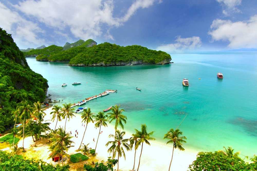 Tropical beach with palm trees, turquoise water, and boats near lush green islands.