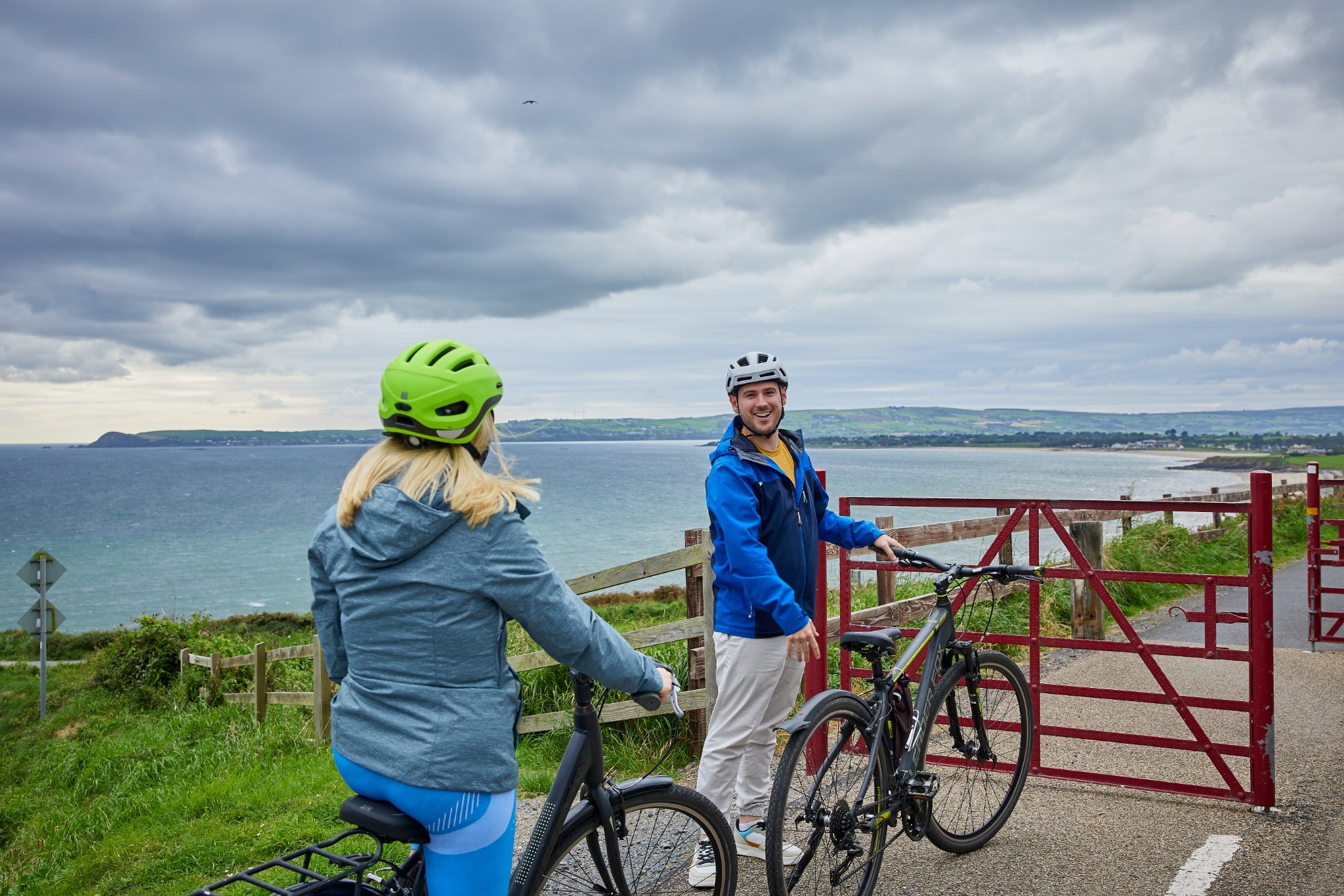 A man and a woman in cycling gear stand with their bikes on a path overlooking the sea.