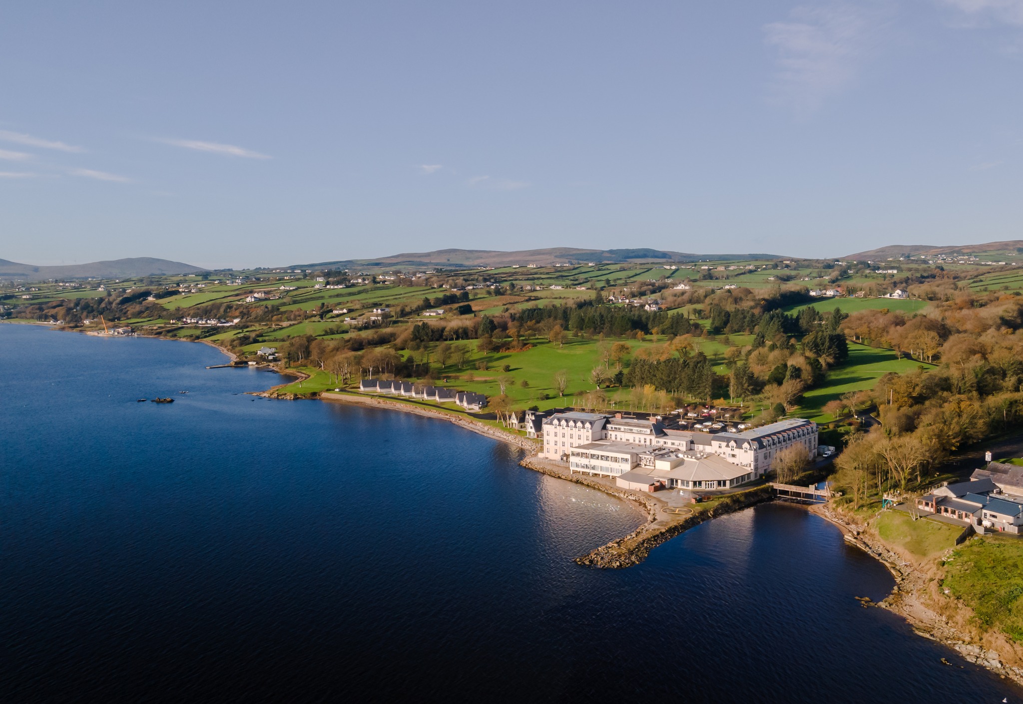 An aerial view of a large hotel on the coast of a bay, with green rolling hills behind.