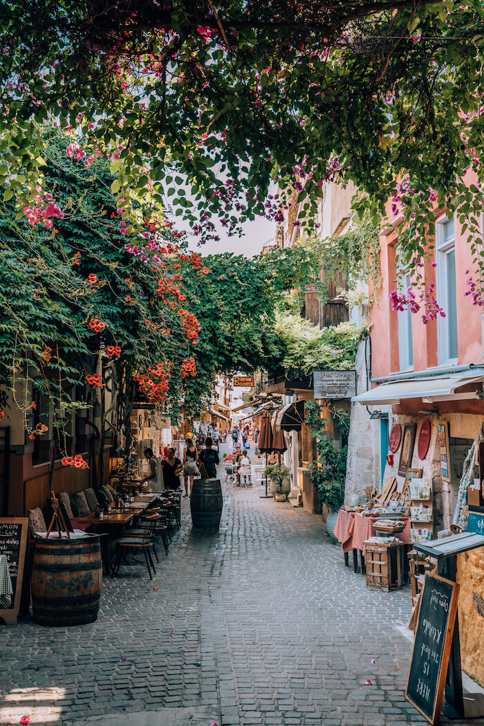Charming cobblestone street with outdoor cafes, shops, and lush hanging flowers.