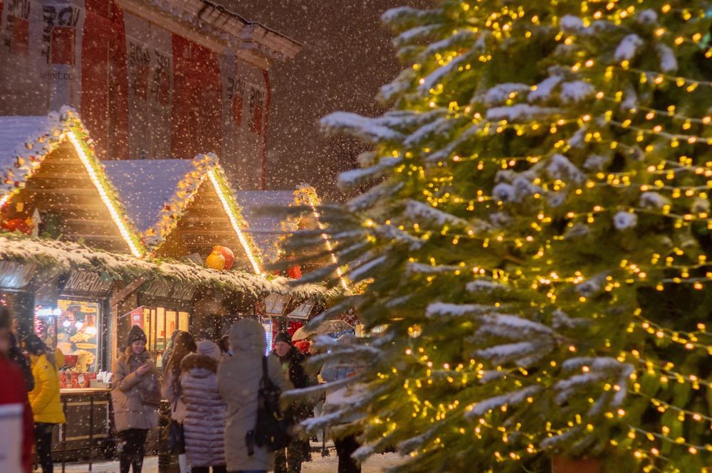 Snowy Christmas market with lit stalls and a decorated tree.