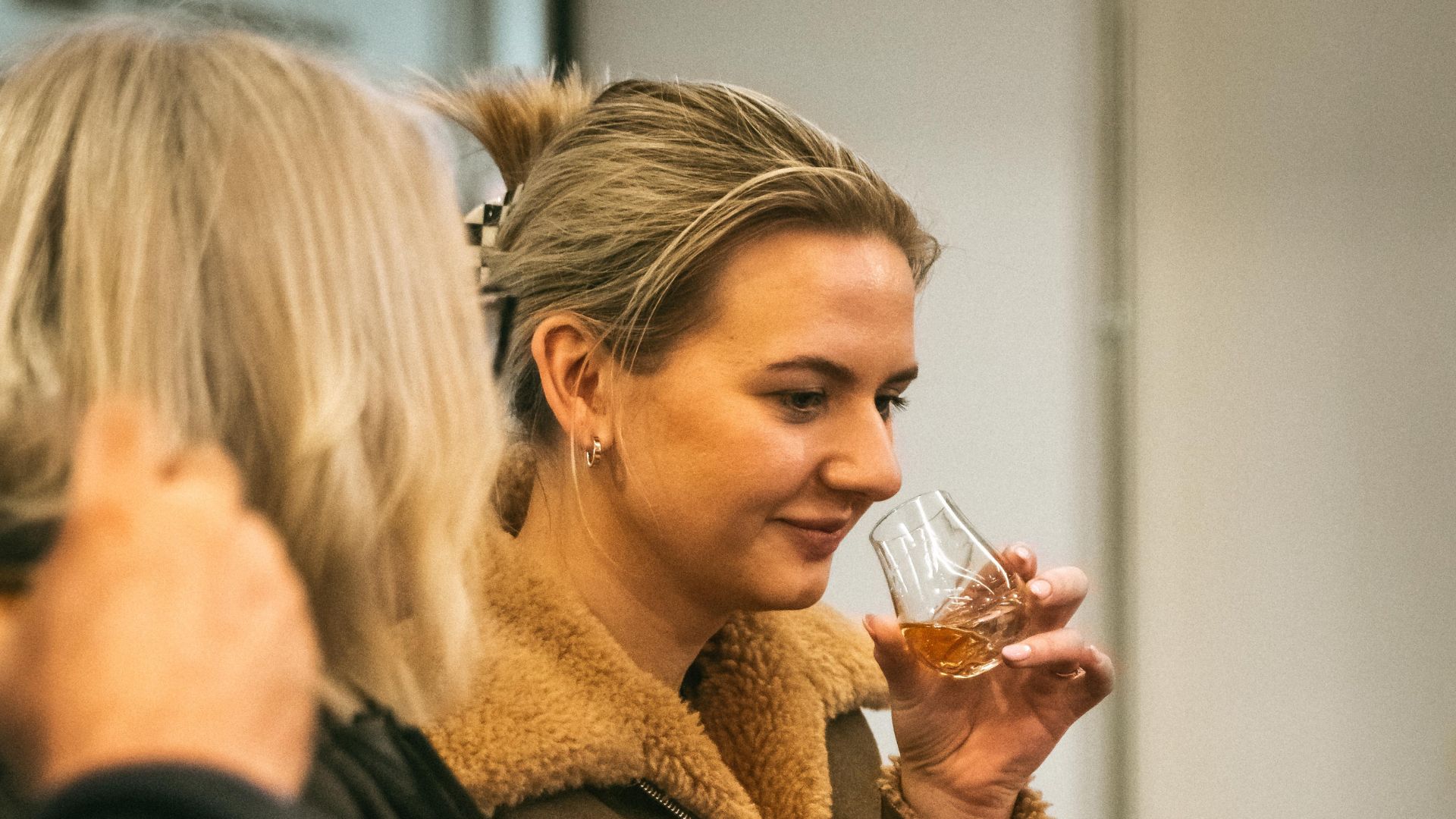 Person smelling a glass of amber liquid, with another person nearby.