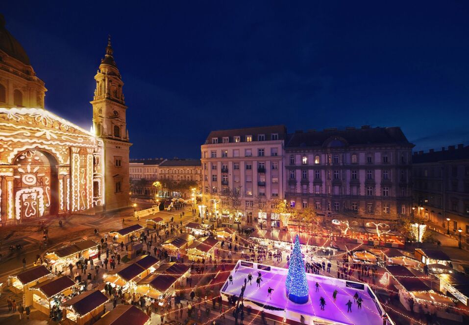An elevated view of a festive Christmas market and ice skating rink in a city square at night.