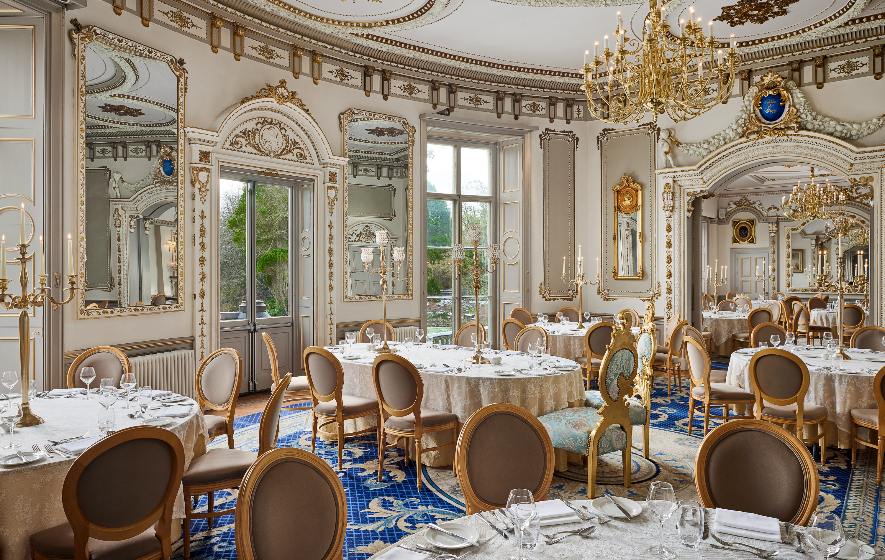Elegant dining room with ornate mirrors, chandeliers, and round tables set for a formal event.